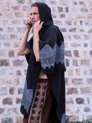 A person wearing the Swing Shawl in black with white geometric patterns stands sideways against a stone wall, adjusting the hood and pairing it with brown pants featuring white designs.
