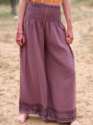 Barefoot on sand, a person wears dusty pink Medusa Palazzo Pants with black embroidered hems and an orange top. Their hands are decorated with intricate henna. The upper body is not shown.