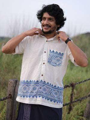 A man stands outdoors smiling, adjusting his Bohemian Tribal Shirt—a cream-colored shirt featuring blue geometric patterns and crafted from breathable cotton—against a blurred background of greenery and a wooden fence.