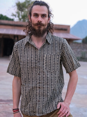 A man with a beard and long curly hair stands outdoors, wearing the Tribal Triangle shirt in black. Blurred buildings and trees can be seen in the background.