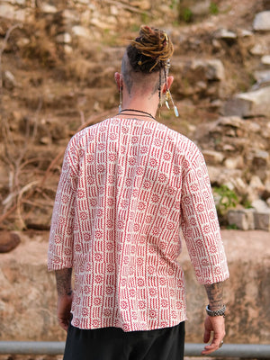 A person with dreadlocks and beaded hair accessories, seen from behind outdoors amid dry foliage and rocks, wears the Moche Kurta—a loose, hand block printed 100% cotton piece with red and white geometric designs—paired with black pants.