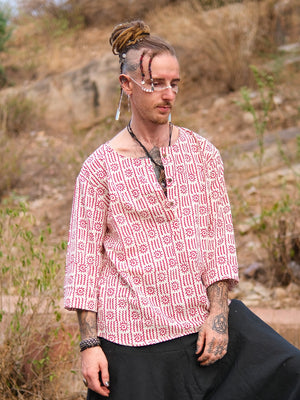 A person with dreadlocks, facial jewelry, and tattoos wears a hand block printed red and white Moche Kurta made from 100% cotton with black pants, standing outdoors in a dry, grassy area with rocks behind them.