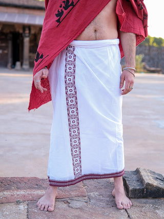 A person stands outdoors in the Chakrot Dhoti, featuring a white wrap with an ornate border around the waist and a stylish red draped cloth over one shoulder. Their bare feet, beautifully enhanced with silver rings and bracelets, complement the high-quality fabric.