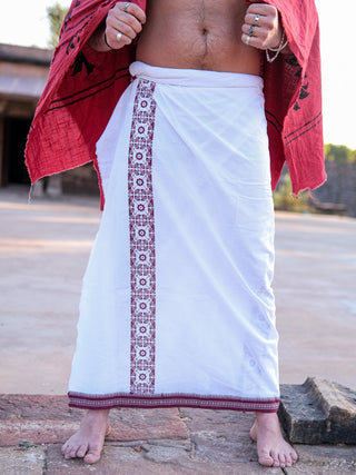 Outdoors, a person stands on a stone surface wearing a stylish red Chakrot Dhoti with maroon border embroidery, adorned with rings on their fingers and draped in a red shawl.