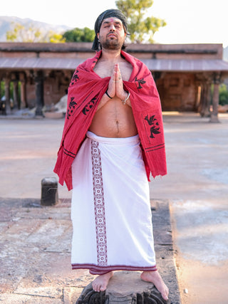A man, eyes closed in prayer, is draped in a red shawl and wearing a pristine Chakrot Dhoti. He stands outdoors against a rustic building surrounded by lush greenery, enjoying the tranquility of daytime.