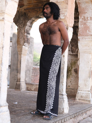 A man with curly hair stands barefoot in a stone archway, wearing the Jasper Dhoti—a black garment featuring white geometric patterns. He is shirtless and gazes off to the side, framed by weathered stone columns and walls.