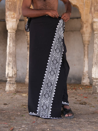 A person wears the Jasper Dhoti in black, featuring white geometric patterns, and adjusts it at the waist. Bare-chested and wearing sandals, they stand against stone columns and an old stone floor.