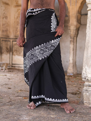 A man stands barefoot, wearing the Jasper Dhoti—a black fabric with white geometric block prints—wrapped around his waist and lower body, set against an old arched stone backdrop. His upper body is not visible.