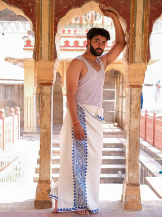 A man stands barefoot in an ornate historic corridor, wearing the Jasper Dhoti with blue block print patterns and a white tank top, resting one arm on an arched pillar while looking at the camera.
