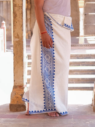 A person stands barefoot in a sunlit, rustic stone corridor, wearing the Jasper Dhoti—white, draped like a sarong, featuring blue geometric border patterns.