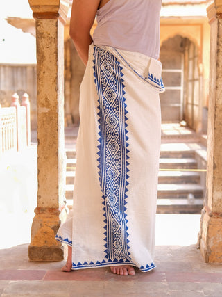 Someone stands barefoot on a stone floor near weathered columns, facing away from the camera while wearing the Jasper Dhoti in white with blue geometric block print patterns.