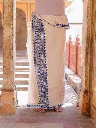 A person stands barefoot in a white Jasper Dhoti with blue geometric patterns, likely block printed, set in a sunlit historic location featuring elegant columns and archways.