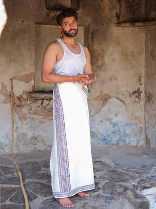 A man with a beard stands barefoot on a stone floor, dressed in a white sleeveless vest and the Nilaya Dhoti, made from premium cotton with a subtle block print, in front of a rustic weathered wall.