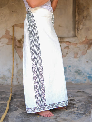 A person stands barefoot on a stone floor, wearing the Nilaya Dhoti—a long white skirt with black block print design borders, made from premium cotton—set against a rustic, textured wall.