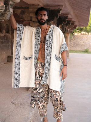 A man wears the Neer Poncho, a cream-colored block print garment with black and white geometric patterned borders and matching pockets, paired with patterned drop-crotch trousers.