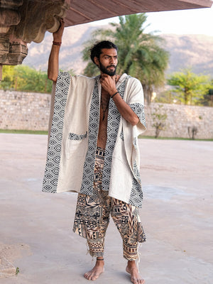 A man stands under an outdoor stone structure wearing an open, boho-style beige Neer Poncho with a black-and-white geometric border and matching patterned drop-crotch pants.