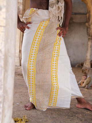 A person stands barefoot in a white and yellow Nira Dhoti made from premium fabric, with a scarf draped over their shoulder. Weathered stone pillars and walls form the background; the persons face remains unseen.