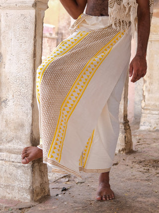 A person wearing the Nira Dhoti, featuring traditional white fabric with yellow and brown block prints, stands barefoot and leans against a weathered pillar, with only their lower body visible.