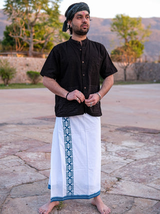 A person stands outdoors on a stone surface, wearing a black shirt and the stylish Chakrot Dhoti made of high-quality fabric with blue patterns. They have a black headwrap and a red mark on their forehead, with trees and a mountain in the background.