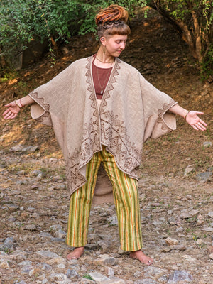 A person with dreadlocks stands barefoot on rocky ground outdoors, wearing the Miraya Shrug over a brown shirt and green-yellow striped pants, arms outstretched with trees in the background.