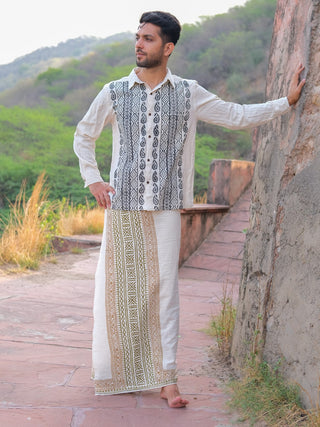 A man stands barefoot outside on a stone path, wearing the Chetra Dhoti—white with black and gold patterns—while resting one arm on a stone wall, with greenery and hills in the background.