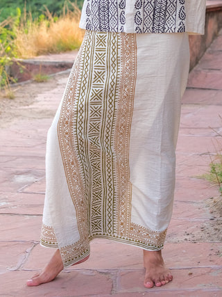 A person stands barefoot on a stone path, wearing a cream Chetra Dhoti with gold and black designs—a timeless element of Indian attire. The scene features grass and a stone wall in the background.
