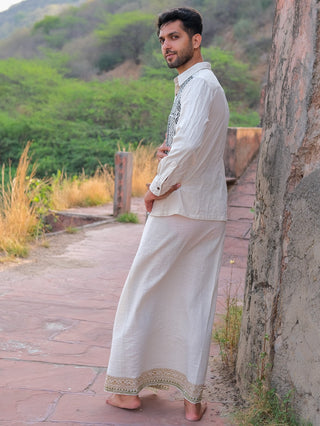 A man models the Chetra Dhoti, standing barefoot on a stone pathway surrounded by greenery and hills, gazing over his shoulder toward the camera.