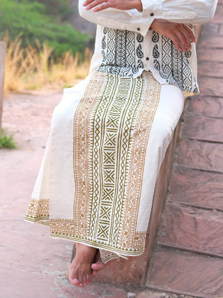 A person sits on a stone bench in a Chetra Dhoti, its intricate gold and black patterns standing out. Barefoot, with hands on their lap, they are surrounded by softly blurred lush greenery from the traditional Indian wardrobe.