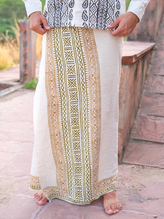 A person stands barefoot on a stone path, wearing a natural Chetra Dhoti and a patterned shirt. The image is cropped to show only the lower body.