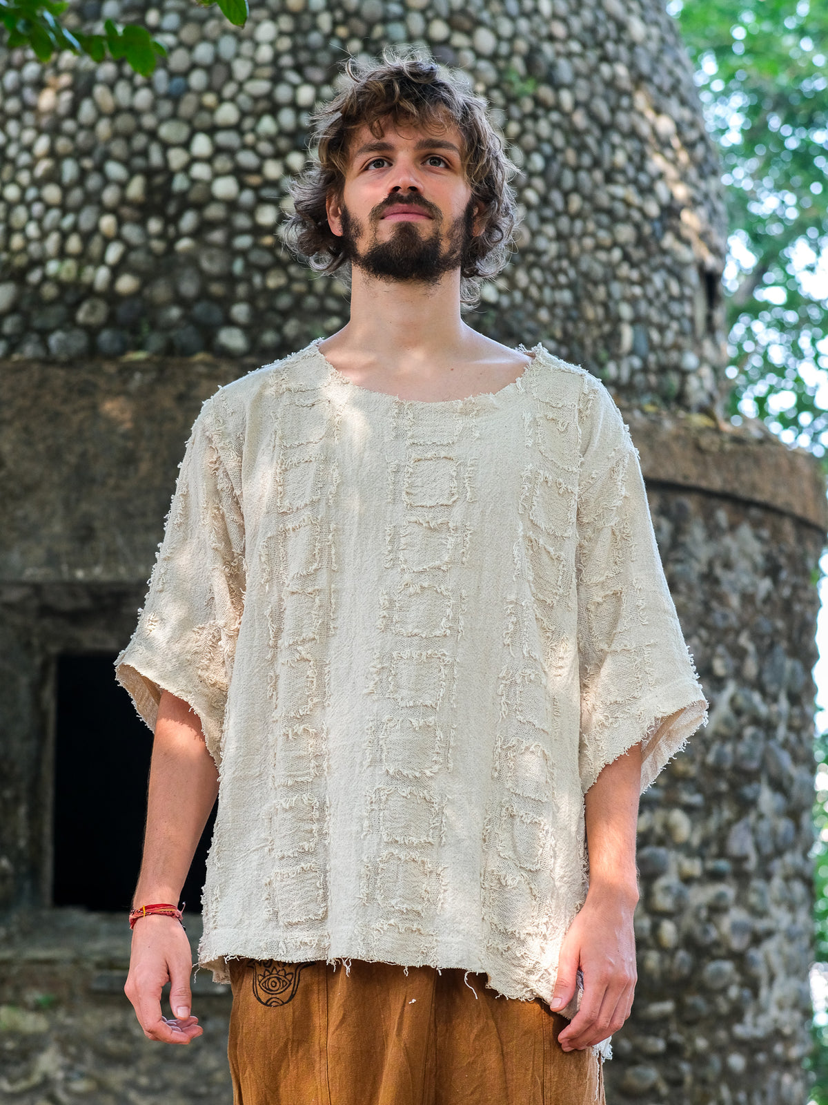 A man with curly hair and a beard stands outside a round stone building in the cream-colored Lennon T-shirt and brown pants, sunlight highlighting his relaxed, bohemian look.