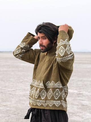 In a spacious, empty landscape, a person with windswept dark hair adjusts the hood of their Tribal Hoodie. Its green fabric is decorated with white geometric patterns against the wide expanse of pale earth under a light sky.