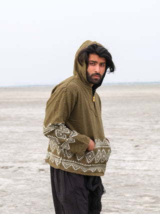 A person with long hair stands in a vast, empty landscape, wearing a Tribal Hoodie with white patterns and black pants. Their hands are tucked into pockets as the sky is overcast, and the ground is barren.