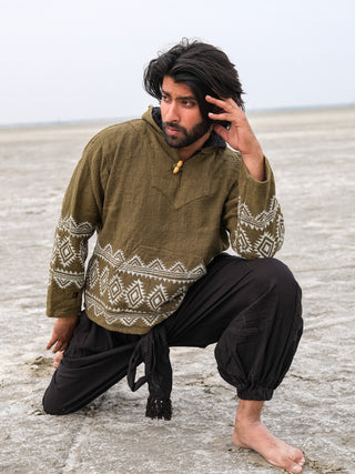 A long-haired, bearded man poses dramatically on sand beneath an overcast sky. He wears the Tribal Hoodie featuring a green block print with white geometric patterns, black pants, and is barefoot. His left hand touches his face while the other rests on his knee.