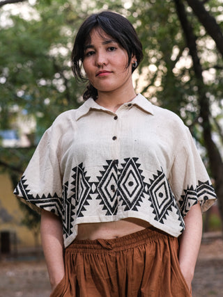 A person with long dark hair stands outdoors, wearing a short-sleeved HWC Tribal Crop Shirt paired with brown pants. The backdrop shows blurred greenery and a muted building.