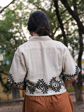 A person with long dark hair, wearing an HWC Tribal Crop Shirt and a brown skirt, is standing outdoors with trees and greenery creating a lush backdrop.