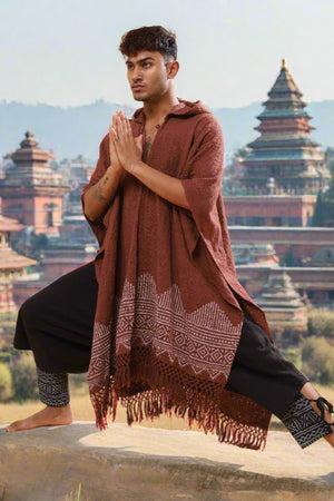 Wearing the brown Swing Shawl and black pants, a person does a yoga pose with hands in prayer against a backdrop of traditional pagoda buildings and distant hills.