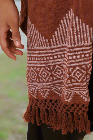 A hand is next to the Swing Shawl, a brown woven traditional shawl with white geometric patterns and tassels at the bottom. The background is blurred green and light brown.