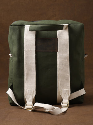 Introducing the Kelly Back Pack—a stylish green canvas backpack featuring crisp white straps and a distinctive brown leather patch, beautifully set against a brown backdrop. This versatile pack is the perfect companion for all your adventures.