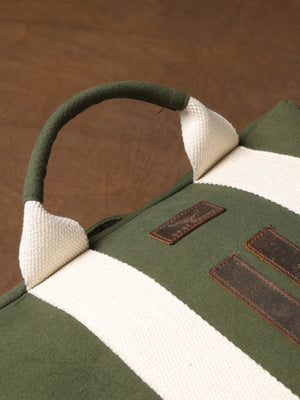 A close-up of the Kelly Back Pack—a green and white striped fabric bag featuring a sturdy handle and two brown leather patches—rests on a brown surface, highlighting this stylish backpack as the perfect adventure companion.