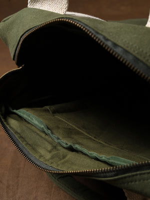 The Kelly Back Pack, an open olive green fabric bag featuring a visible zipper, white handles, and a roomy inner compartment, is displayed on a brown surface—making it the perfect companion for any adventure.
