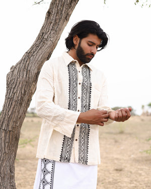 A man with medium-length dark hair and a beard adjusts his sleeve while wearing the Acharya Shirt Full-sleeve, a cream and black embroidered minimalistic design, next to a tree in a grassy field under a cloudy sky.