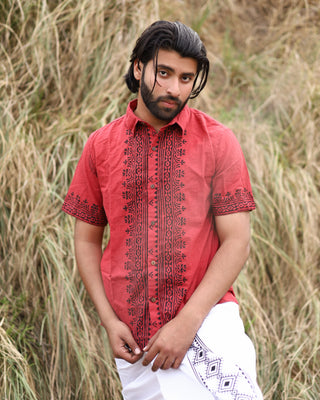 A man with medium dark hair and a beard stands outdoors by tall dry grass, wearing the eco-friendly red Likhit Shirt, blockprinted by hand with black patterns, and a white garment wrapped around his waist.