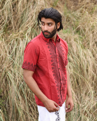 A man with dark hair and a beard stands outdoors before tall dry grass, looking right. He wears the Likhit Shirt—a red, hand block-printed shirt—and white pants.
