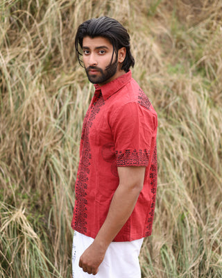 A dark-haired, bearded man wears the red Likhit Shirt, featuring hand block-printed black patterns, paired with white pants and standing outdoors before tall dry grass while looking at the camera.