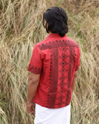 A person with medium-length dark hair stands outdoors with their back to the camera, wearing a red Likhit Shirt featuring hand-blockprinted black geometric and floral designs, paired with white pants amid tall dry grass.