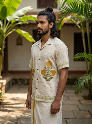 A man with long hair in a bun stands outdoors, wearing the Sunehri Shirt—a hand block printed cream shirt adorned with yellow flowers—paired with a light traditional garment. Lush green plants and a building can be seen in the background.