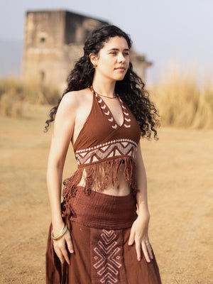 A woman with curly hair wearing the Divine Crop Top and a matching skirt stands outdoors on dry grass, with an old stone building blurred in the background.