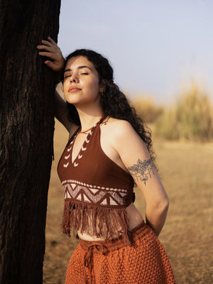 A woman with curly hair leans against a tree outdoors, wearing the Divine Crop Top—a boho-inspired piece featuring a subtle block print—paired with an orange skirt. Sunlight illuminates her face as her floral tattoo is visible on her upper arm.