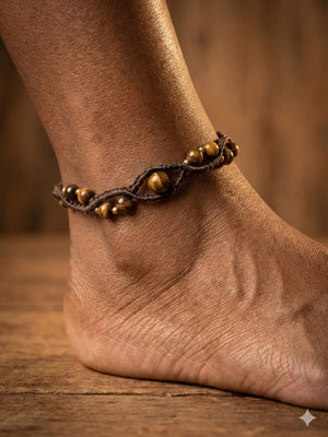 A close-up of an ankle adorned with the Aarohi Aventurine Boho Anklet in brown, featuring a braided macramé design with gold-colored beads, set against a wooden background.