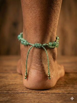 A close-up of a person’s ankle wearing the Aarohi Aventurine Boho Anklet, featuring green beads and dangling strings, standing on a wooden floor with a blurred brown background.